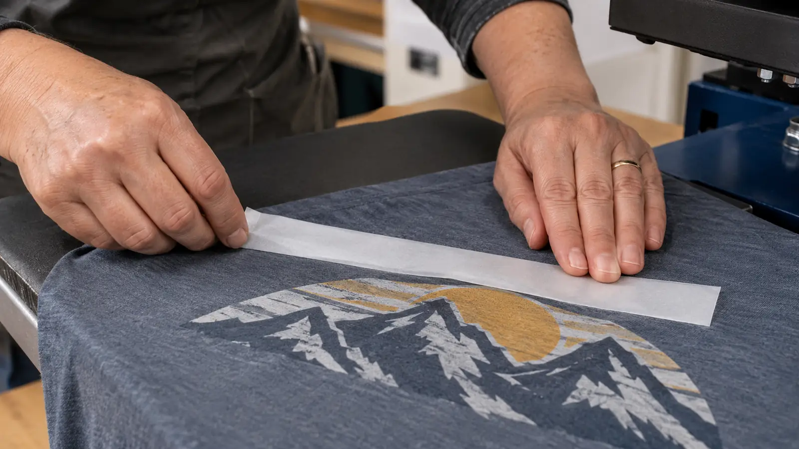 A hand pressing a strip of heat tape over the edge of a transfer on a shirt before pressing